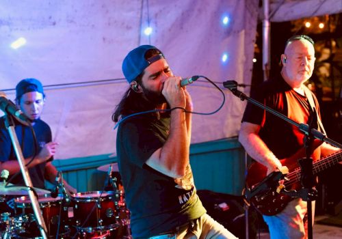 A band is performing on stage with a drummer, vocalist, and bassist, under colorful lights and a tent.