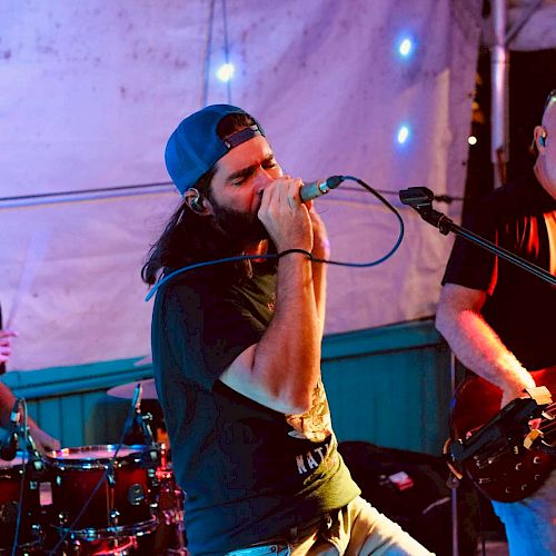 A band is performing on stage with a drummer, vocalist, and bassist, under colorful lights and a tent.