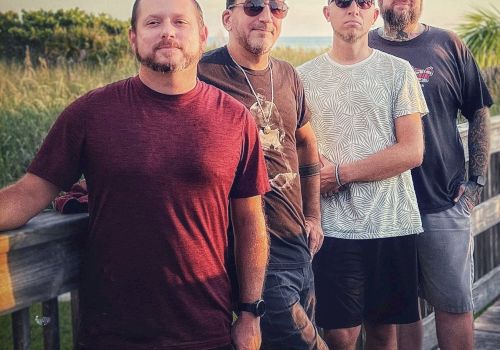 This image features four men standing on a wooden walkway outdoors with text at the top that reads 