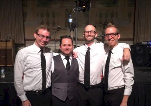 Four people in formal attire are standing together, smiling, possibly part of a band or group at an event.