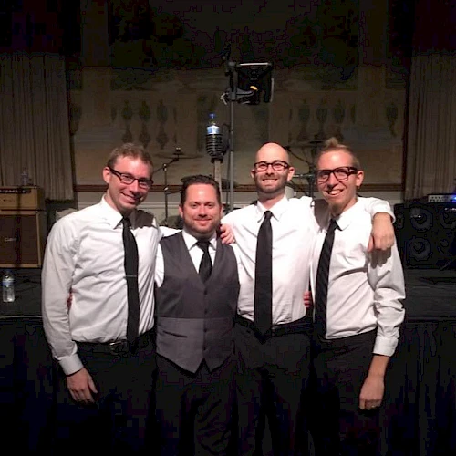Four people in formal attire are standing together, smiling, possibly part of a band or group at an event.