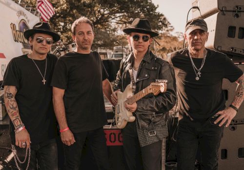 Four men are standing together outdoors, one holding an electric guitar. They all wear dark clothing and accessories, posing for the photo.