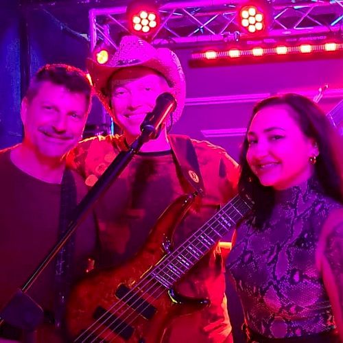 Four people posing on stage with instruments under pink and red lighting, one person with a guitar, another with a double bass, smiling.