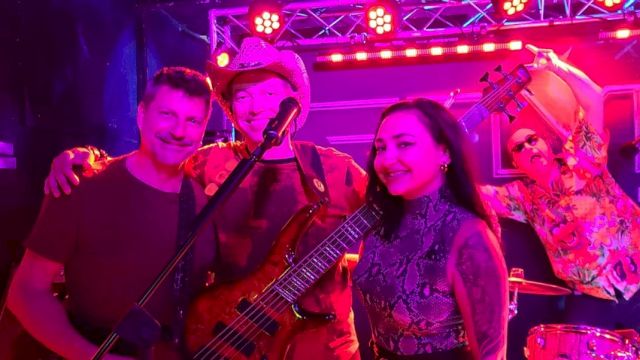 Four people posing on stage with instruments under pink and red lighting, one person with a guitar, another with a double bass, smiling.
