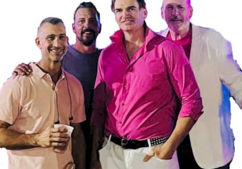 The image shows four men standing together, smiling for the camera. The man in front is wearing a bright pink shirt with white pants.