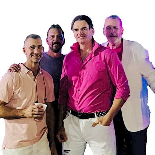 The image shows four men standing together, smiling for the camera. The man in front is wearing a bright pink shirt with white pants.