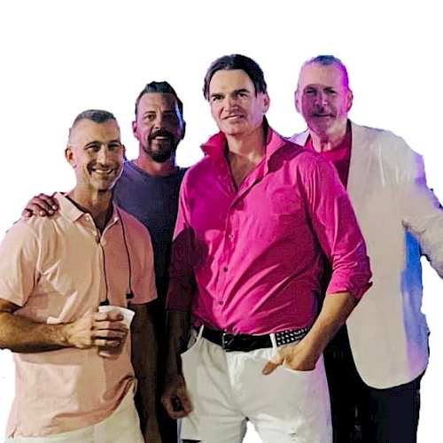 The image shows four men standing together, smiling for the camera. The man in front is wearing a bright pink shirt with white pants.