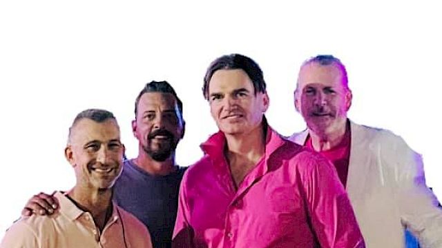 The image shows four men standing together, smiling for the camera. The man in front is wearing a bright pink shirt with white pants.