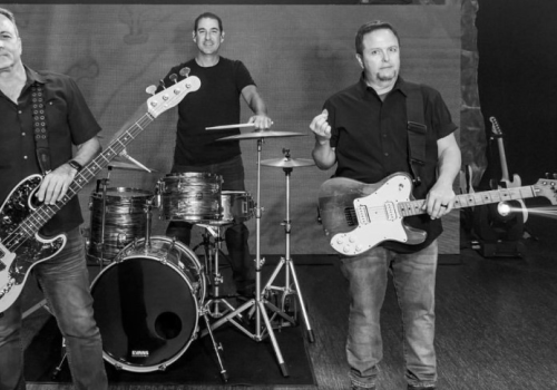 Three men are posing with musical instruments: a bass guitar, a drum set, and an electric guitar, on a stage.