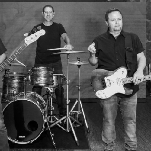 Three men are posing with musical instruments: a bass guitar, a drum set, and an electric guitar, on a stage.