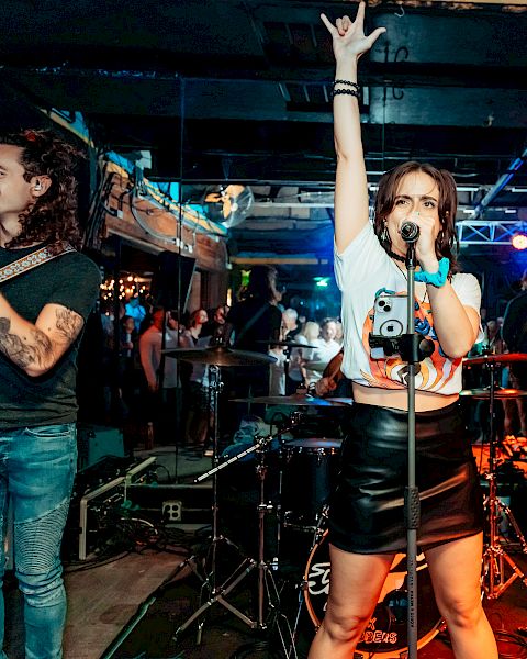 A band performing on stage with a lead singer raising her hand, flanked by guitarists, in a lively venue with colorful lights and people in the background.