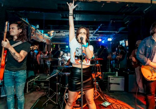 A band performing on stage with a lead singer raising her hand, flanked by guitarists, in a lively venue with colorful lights and people in the background.