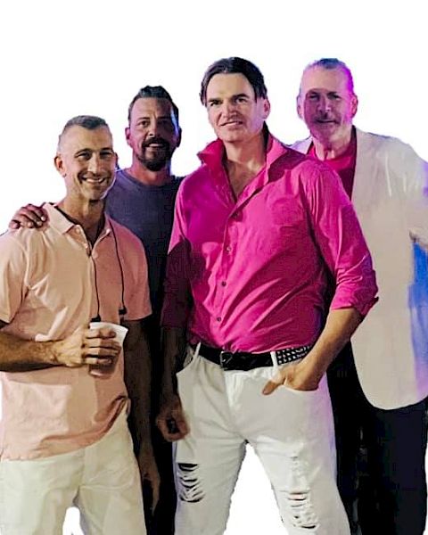 Four men are posing together, three wearing pink shirts and one in a white blazer. The man in front is holding a cup.