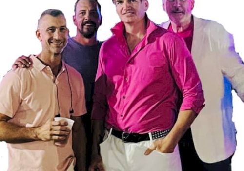 Four men are posing together, three wearing pink shirts and one in a white blazer. The man in front is holding a cup.