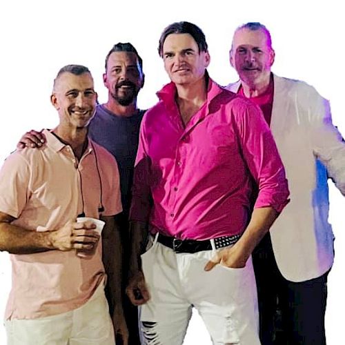 Four men are posing together, three wearing pink shirts and one in a white blazer. The man in front is holding a cup.