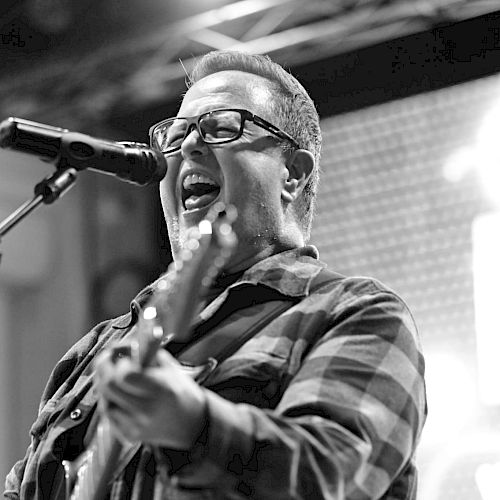 A person passionately singing and playing guitar on stage, wearing glasses and a plaid shirt, captured in black and white.
