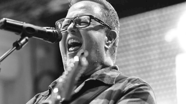 A person passionately singing and playing guitar on stage, wearing glasses and a plaid shirt, captured in black and white.