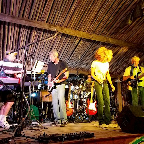 A band performs on a wooden stage under a rustic roof. The members play keyboards, guitars, and sing, with various equipment visible around them.