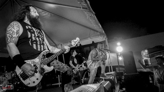 The image shows a black-and-white photo of a band performing on stage, with a guitarist, bassist, and vocalist under a tent.