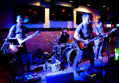 A rock band is performing on stage with members playing guitars, bass, and drums under purple lighting in a venue with a brick wall background.