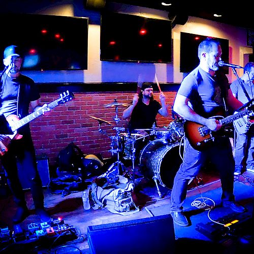 A rock band is performing on stage with members playing guitars, bass, and drums under purple lighting in a venue with a brick wall background.