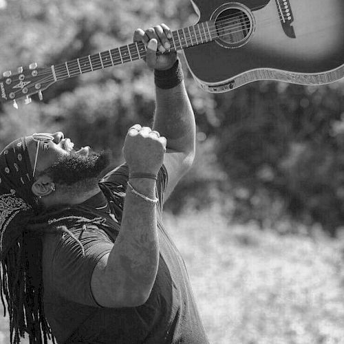 A person energetically raising an acoustic guitar overhead with one hand, while clenching a fist and looking upward.