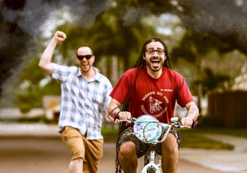 Two men are having fun. One is riding a small bike, and the other is cheering in the background on a street in a casual setting.