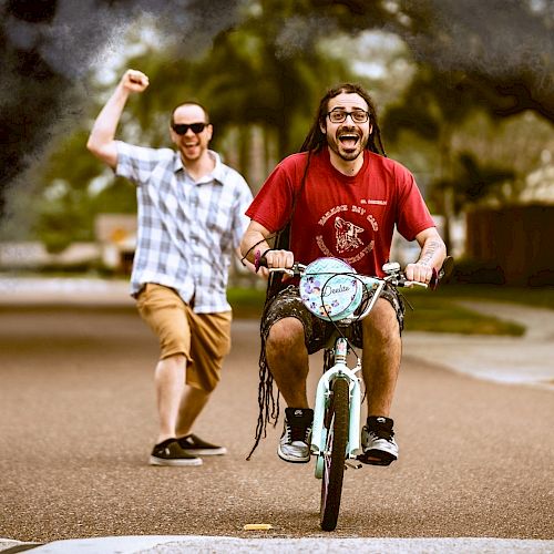 Two men are having fun. One is riding a small bike, and the other is cheering in the background on a street in a casual setting.