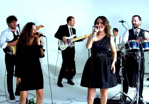 A band is performing in a white studio, featuring guitarists, vocalists, a drummer, a keyboardist, and a percussionist.