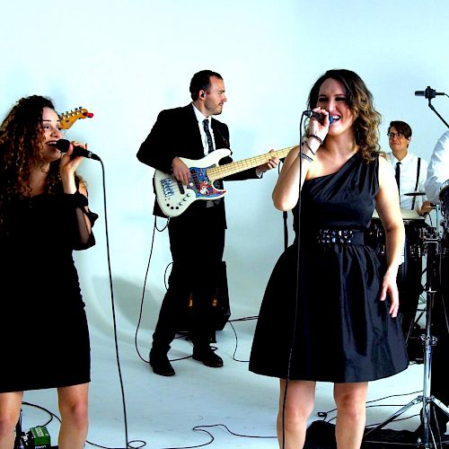 A band is performing in a white studio, featuring guitarists, vocalists, a drummer, a keyboardist, and a percussionist.