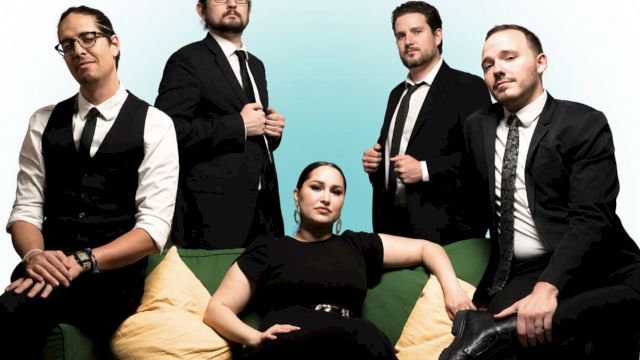 Five people in formal attire, four standing and one seated, posing on and around a green couch with yellow cushions.