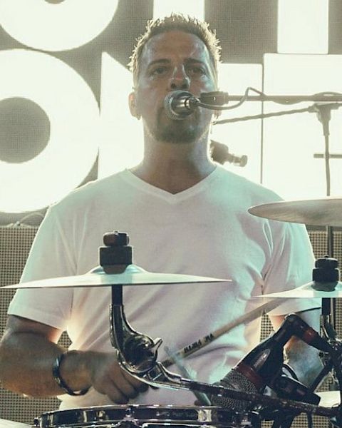 A person is playing drums on stage, wearing a white shirt and singing into a microphone. The background has bright lighting.