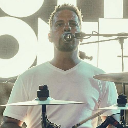 A person is playing drums on stage, wearing a white shirt and singing into a microphone. The background has bright lighting.