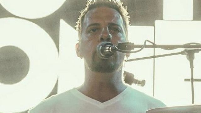 A person is playing drums on stage, wearing a white shirt and singing into a microphone. The background has bright lighting.
