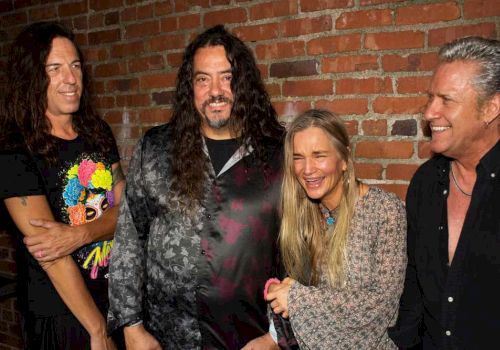 Four people are standing in front of a brick wall, smiling and laughing together.