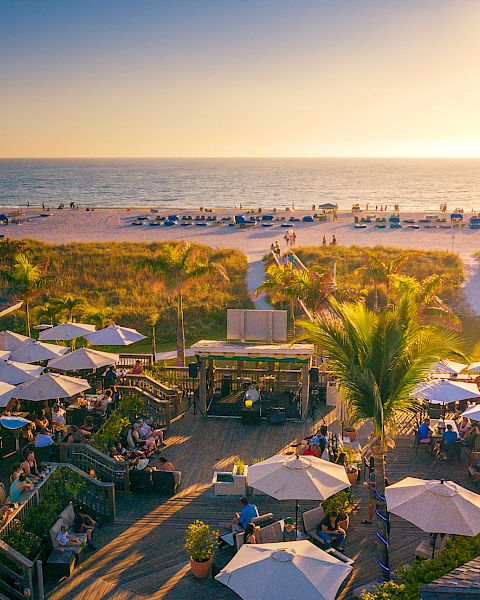 A beachfront scene during sunset with people dining under umbrellas, palm trees, a wooden deck, and the ocean in the background.