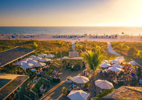 A beachfront scene during sunset with people dining under umbrellas, palm trees, a wooden deck, and the ocean in the background.