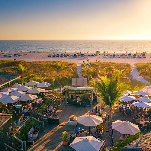 A beachfront scene during sunset with people dining under umbrellas, palm trees, a wooden deck, and the ocean in the background.