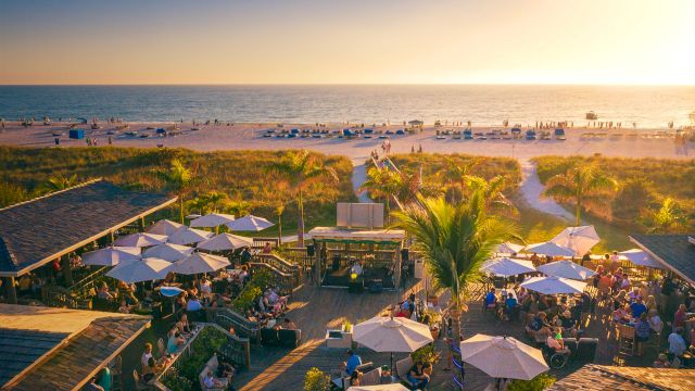 A beachfront scene during sunset with people dining under umbrellas, palm trees, a wooden deck, and the ocean in the background.