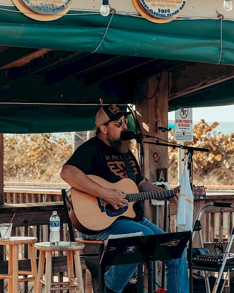 A musician is playing an acoustic guitar and singing on an outdoor stage with a green awning, surrounded by stools and music equipment.