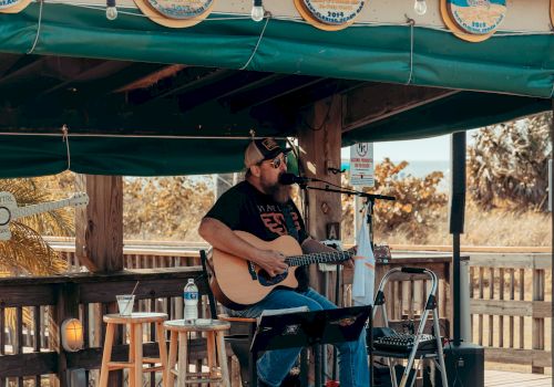 A musician is playing an acoustic guitar and singing on an outdoor stage with a green awning, surrounded by stools and music equipment.