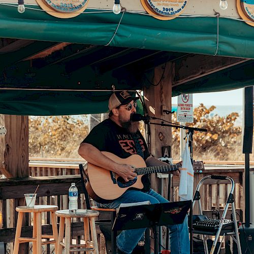 A musician is playing an acoustic guitar and singing on an outdoor stage with a green awning, surrounded by stools and music equipment.