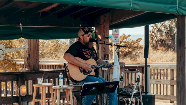 A musician is playing an acoustic guitar and singing on an outdoor stage with a green awning, surrounded by stools and music equipment.