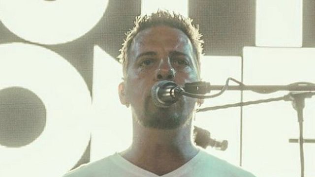 A man in a white shirt is playing the drums and singing into a microphone on stage.