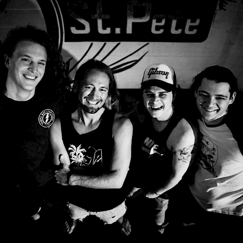 Four people are standing closely together, smiling. The background has a poster with partial text "St. Pete" visible, ending the sentence.