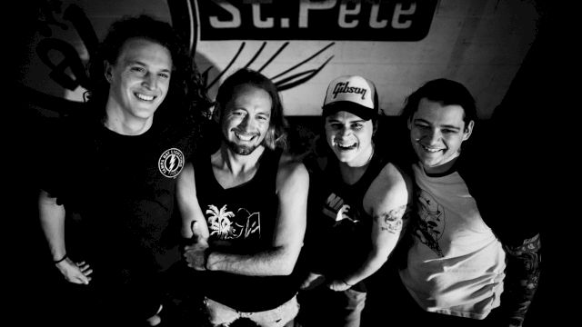 Four people are standing closely together, smiling. The background has a poster with partial text "St. Pete" visible, ending the sentence.