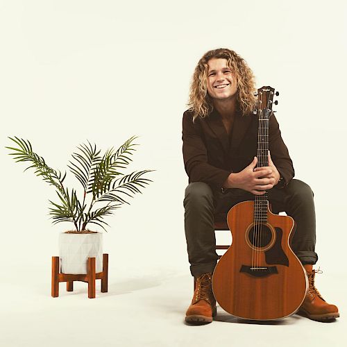 A man with long curly hair is sitting on a chair, holding an acoustic guitar, next to a potted plant in a minimalist setting.