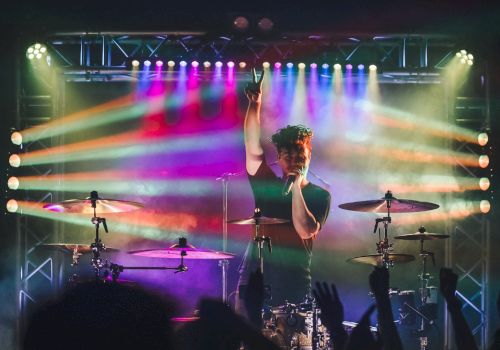 A drummer performs on stage with vibrant colorful lights and a cheering crowd, who raise their hands in appreciation of the music.