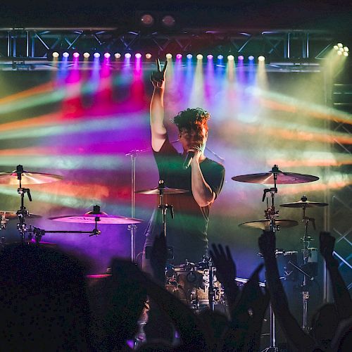 A drummer performs on stage with vibrant colorful lights and a cheering crowd, who raise their hands in appreciation of the music.