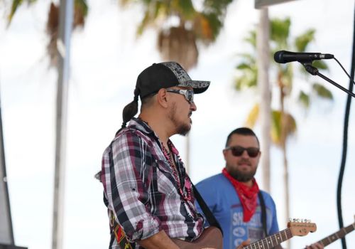 Two musicians are playing electric guitars on an outdoor stage. Palm trees are visible in the background, and they both wear casual clothes and sunglasses.